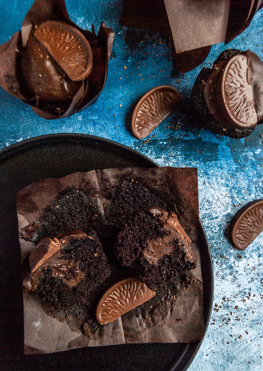 a chocolate muffin on a black plate cut in half to reveal a chocolate orange ganache filling. More chocolate muffins topped with chocolate ganache and a terry's chocolate orange segment sit above the plate.
