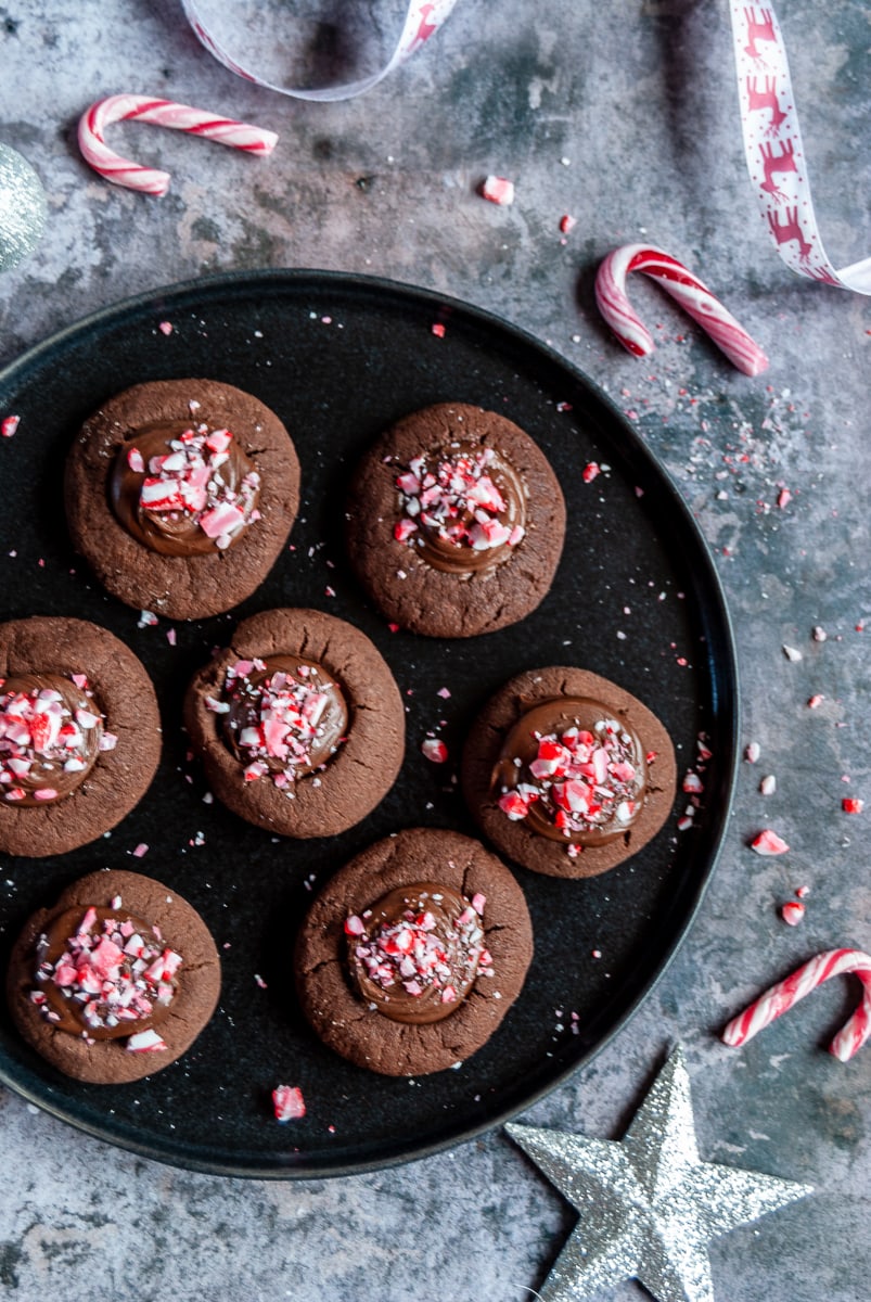 a black plate with chocolate cookies topped with chocolate ganache and crushed peppermint candy canes. mini candy canes and silver christmas decorations have been placed around the plate