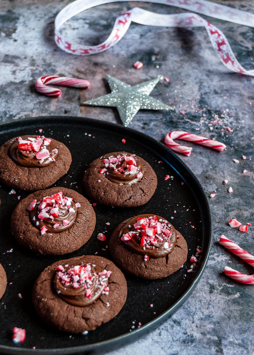 a black plate of chocolate cookies topped with chocolate ganache and sprinkled with crushed peppermint candy canes