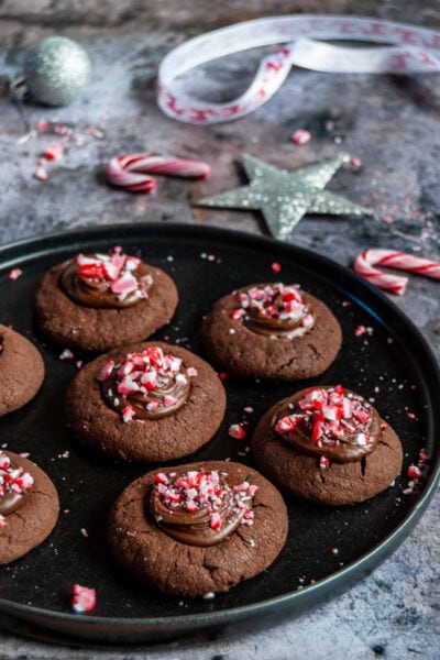a black plate of chocolate cookies topped with chocolate ganache and sprinkled with crushed peppermint candy canes.