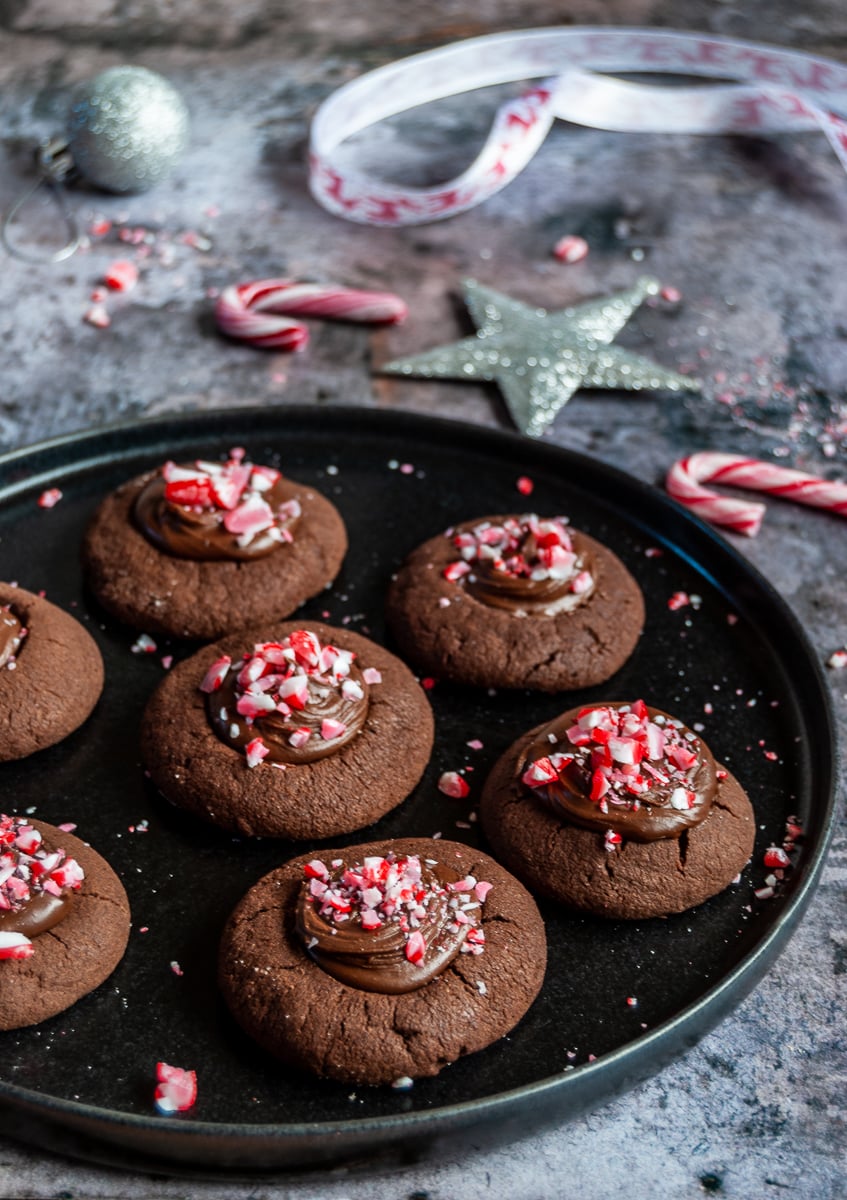 a black plate of chocolate cookies topped with chocolate ganache and sprinkled with crushed peppermint candy canes.