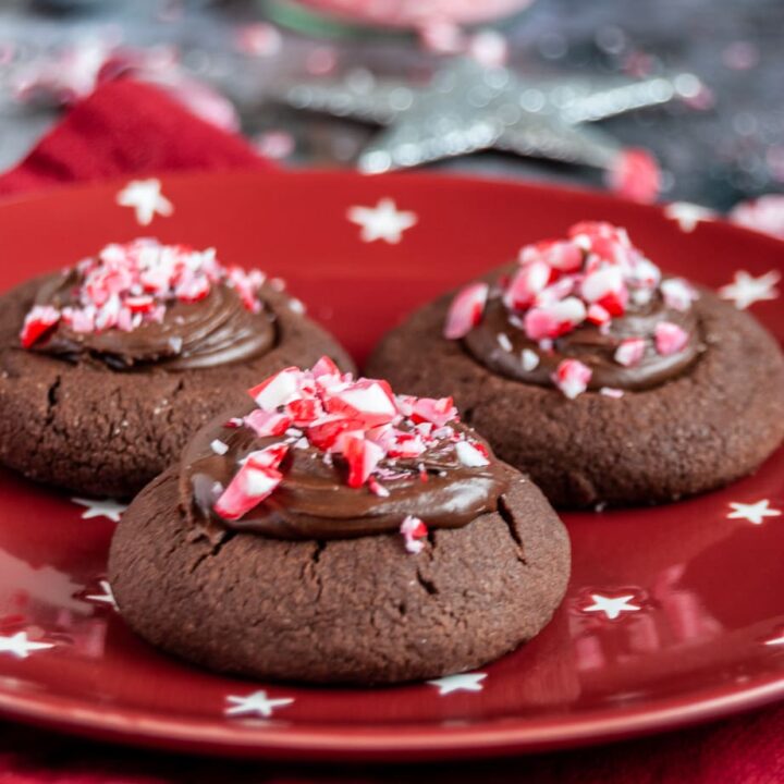 A close up photo of three chocolate cookies topped with chocolate ganache and crushed peppermint candy canes on a red white and star plate and red napkin. Silver Christmas decorations can be partially seen in the background