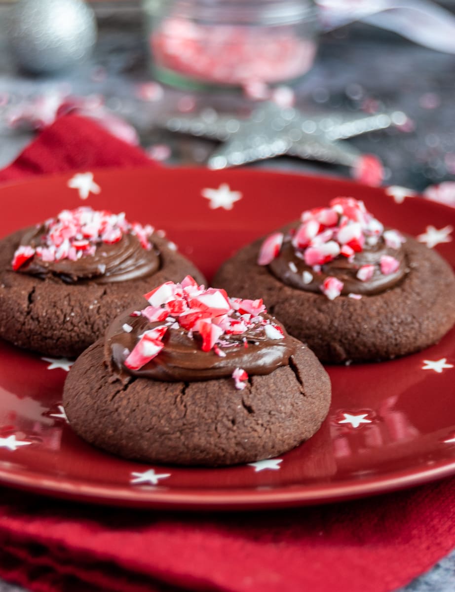 A close up photo of three chocolate cookies topped with chocolate ganache and crushed peppermint candy canes on a red white and star plate and red napkin. Silver Christmas decorations can be partially seen in the background