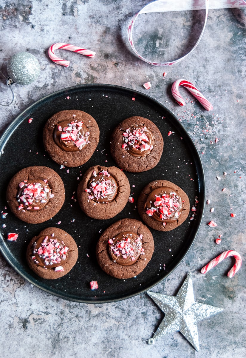 a black plate with chocolate cookies topped with chocolate ganache and crushed peppermint candy canes. mini candy canes and silver christmas decorations have been placed around the plate