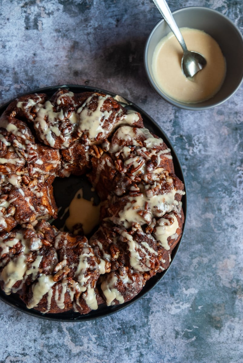 a cinnamon monkey bread with pecans and covered in a maple icing glaze on a black plate, a grey bowl of maple icing and a spoon on a grey background