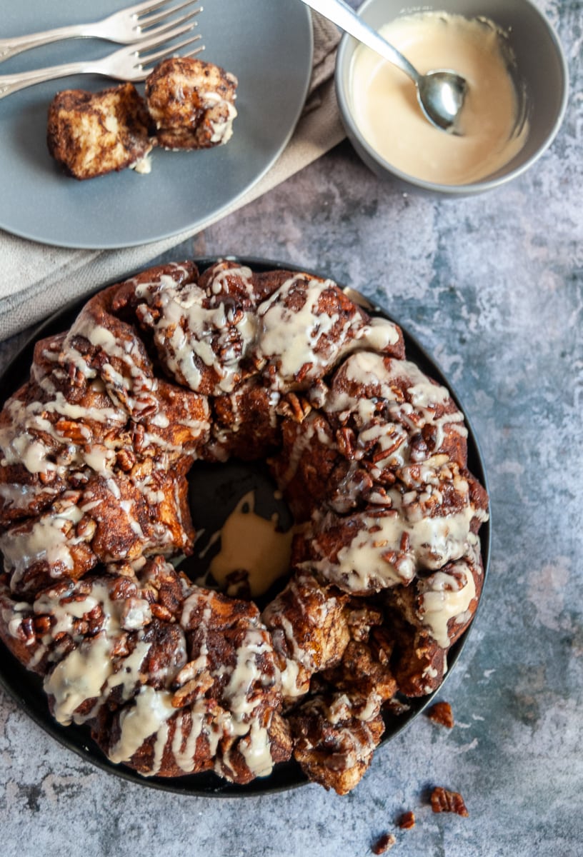 monkey bread with pecans drizzled with a maple glaze on a black plate, a grey bowl of maple icing and a spoon and a grey plate with two pieces of the monkey bread and two forks