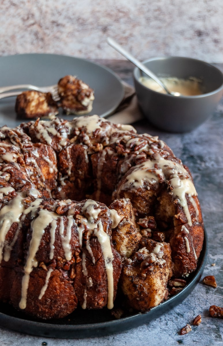 A cinnamon monkey bread stuffed with pecans and covered in a maple icing glaze on a black plate, a grey pot of maple icing with a spoon and two pieces of monkey bread with a fork in the background.
