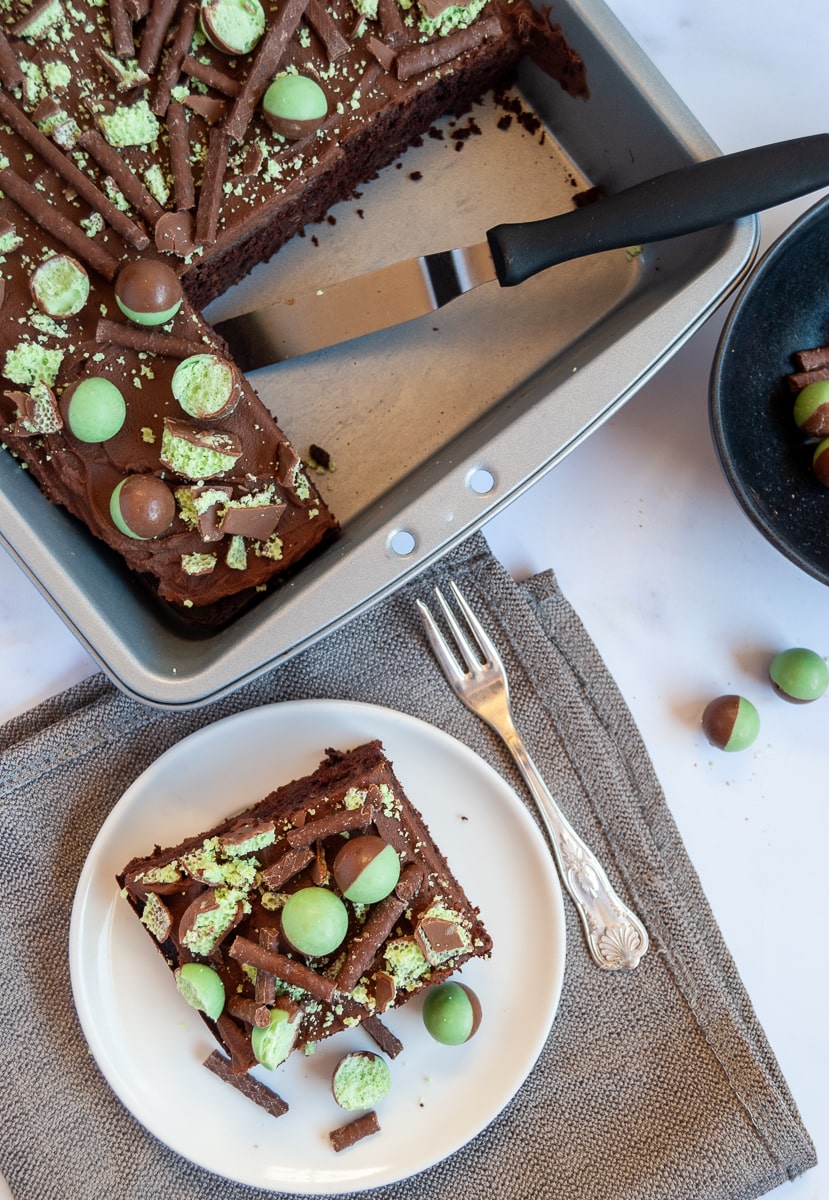 An overhead photo of a chocolate sheet cake topped with chocolate ganache and mint aero bubbles in a baking pan, a white plate with a slice of the cake, a grey napkin and a silver fork.