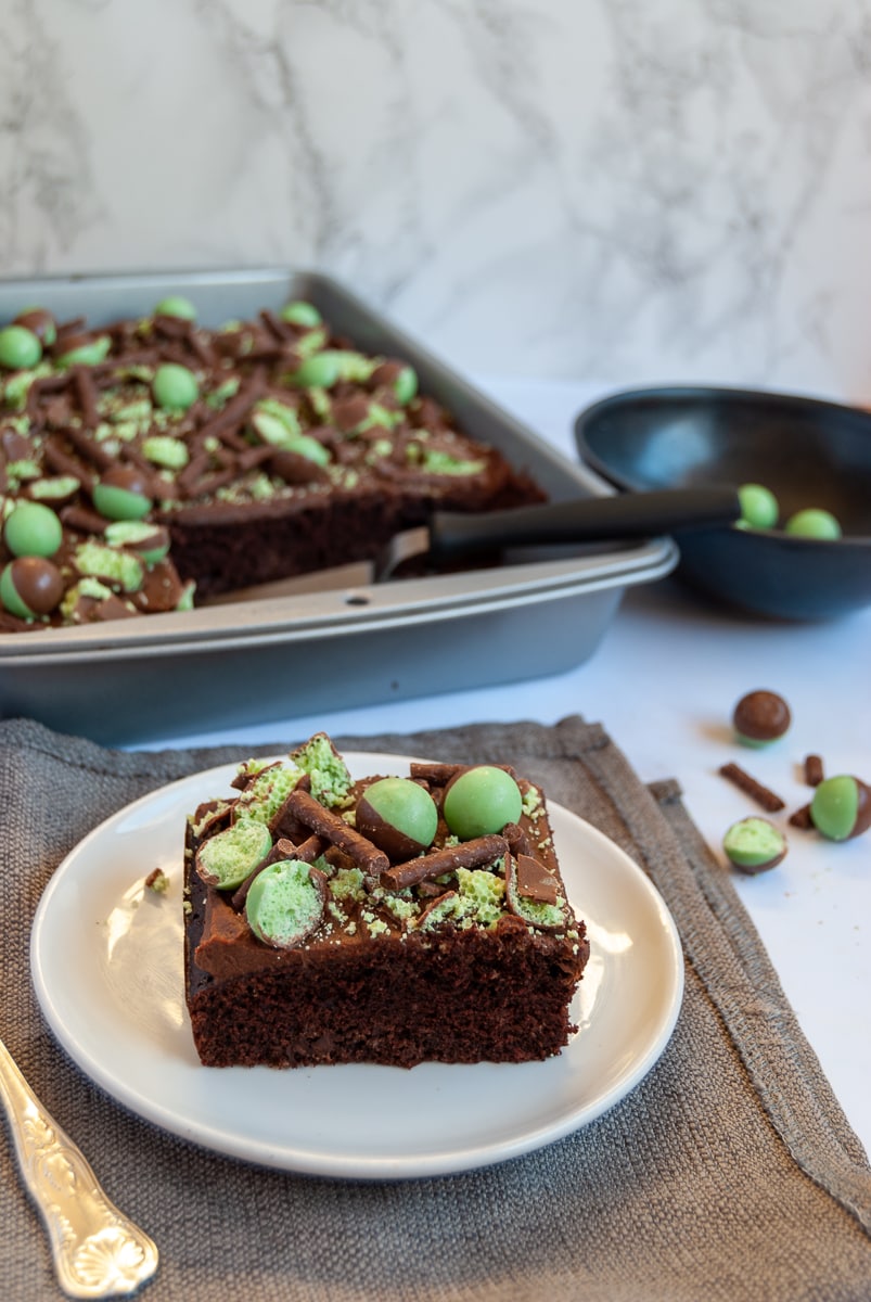 a slice of mint chocolate cake topped with chocolate ganache and mint aero bubbles, a grey napkin and a silver fork. A silver baking pan with more cake can be seen in the background