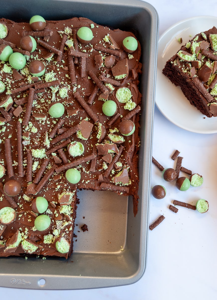 A chocolate sheet cake covered with chocolate ganache and topped with mint aero bubbles in a silver baking pan and a white plate with a piece of chocolate cake