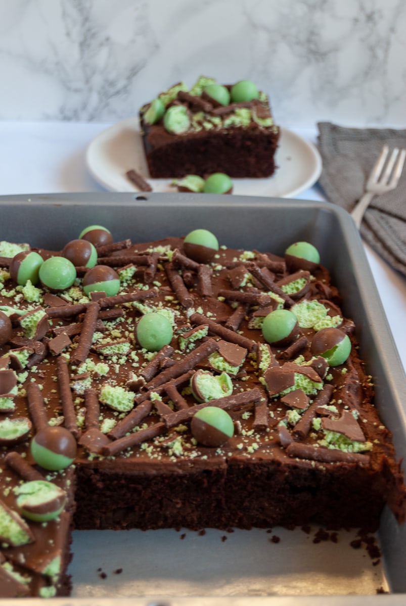 a chocolate sheet cake topped with chocolate ganache and mint Aero bubbles. A white plate with a piece of the cake can be seen in the background