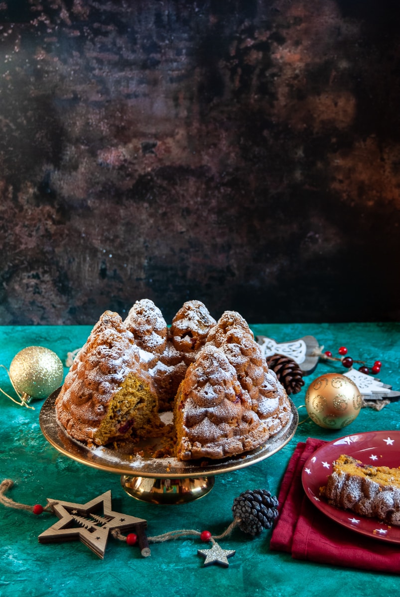 A pumpkin and cranberry bundt cake in the shape of fir trees on a gold cake stand surrounded by christmas decorations on a green and brown backdrop