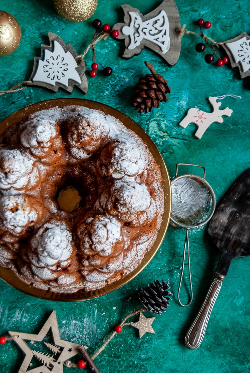 a flat lay photo of a pumpkin bundt cake in the shape of fir trees on a gold cake stand, surrounded by Christmas decorations, a cake slicer and a sugar sifter on a green background