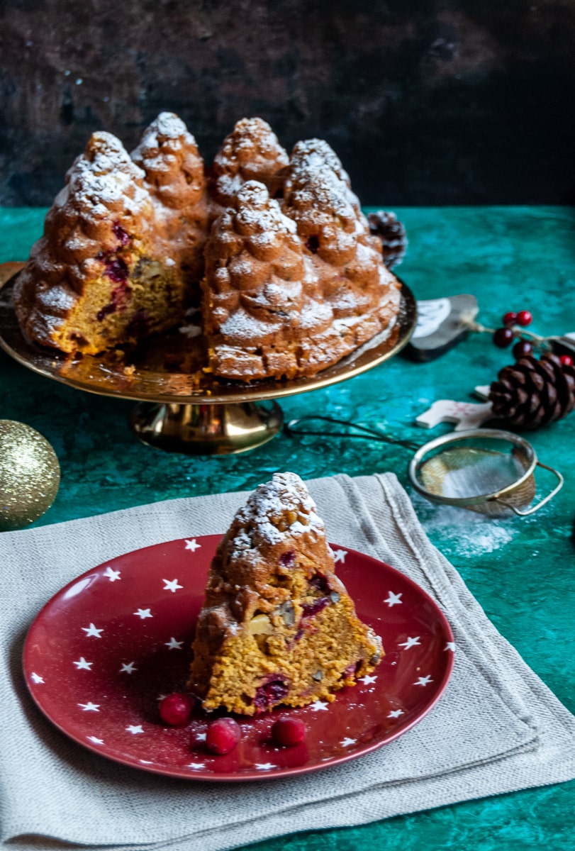 a photo of a slice of pumpkin cake with cranberries and chopped apples on a red and white star plate.  A gold cake stand with the bundt cake can be seen in the background along with Christmas decorations and a small sieve with icing sugar.