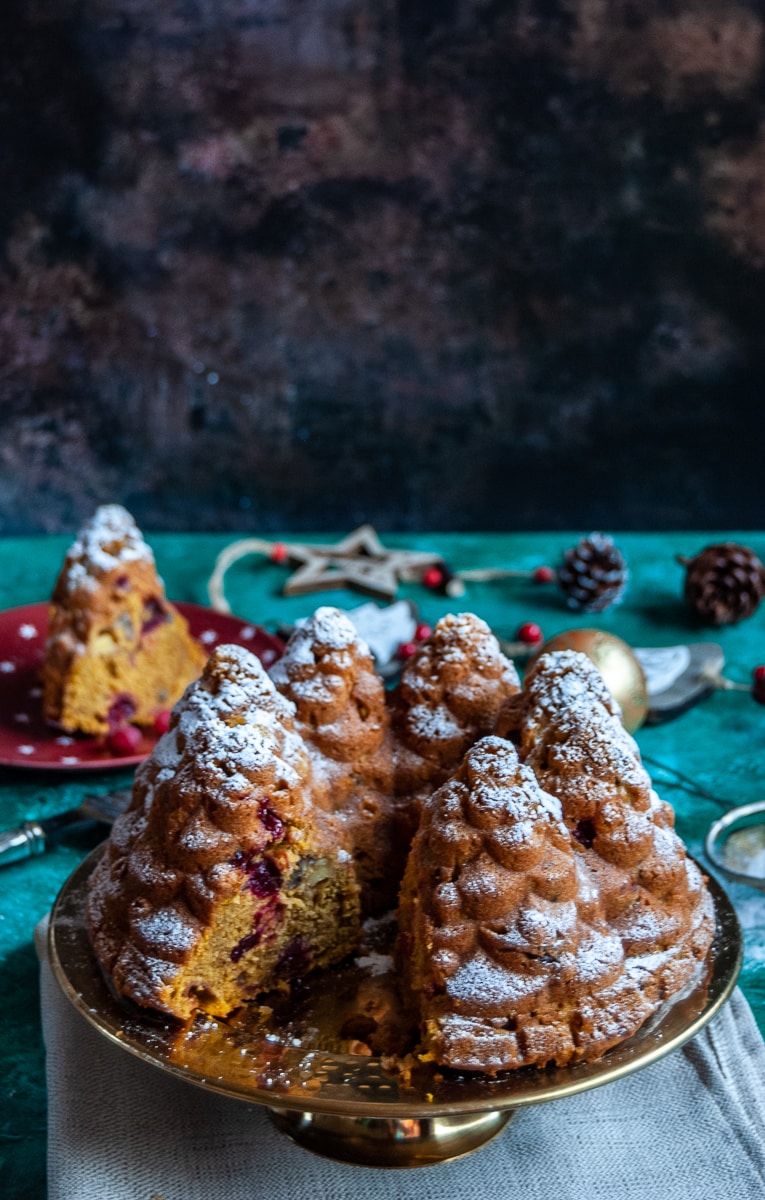 a pumpkin and cranberry bundt cake in the shape of fir trees dusted with icing sugar on a gold cake stand with a slice cut out of it, a red plate with a slice of the cake in the background and christmas decorations