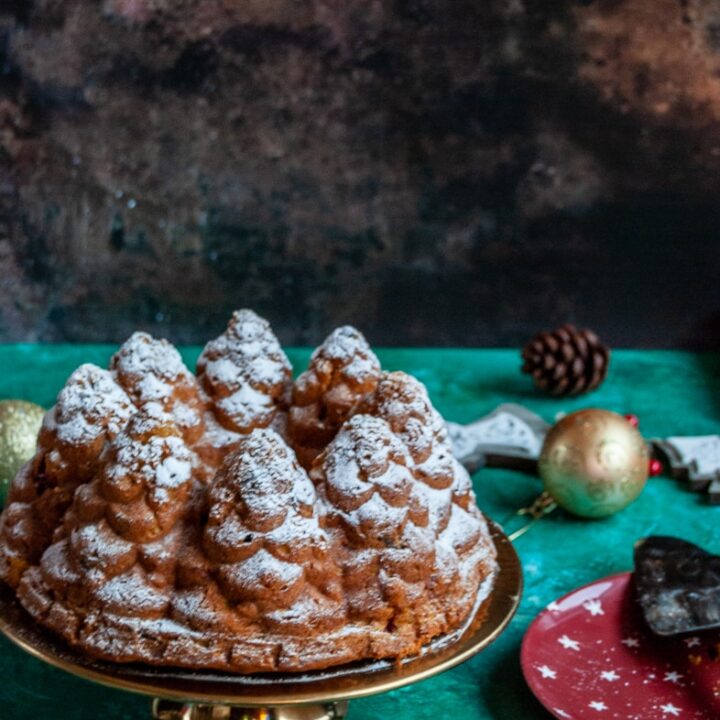 a pumpin bundt cake in the shape of fir trees dusted with icing sugar on a gold cake stand, a red plate with a vintage cake slice and christmas decorations on a brown and green backdrop