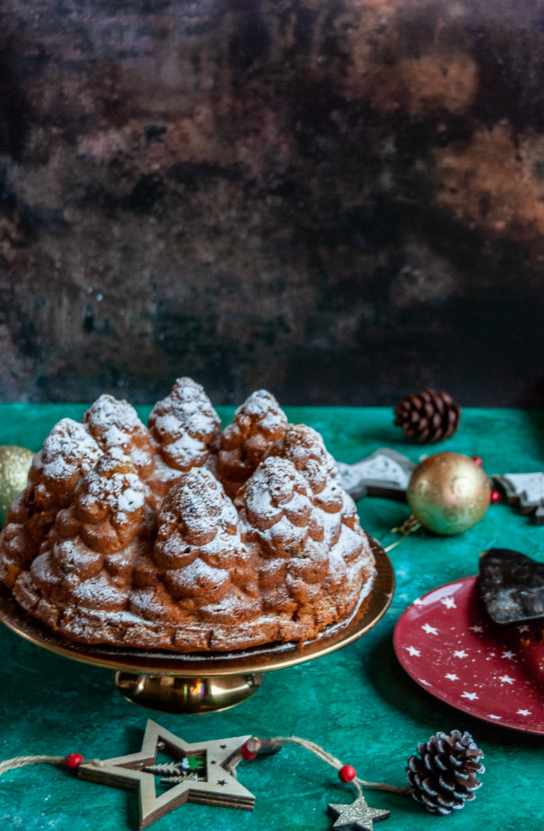 a pumpin bundt cake in the shape of fir trees dusted with icing sugar on a gold cake stand, a red plate with a vintage cake slice and christmas decorations on a brown and green backdrop