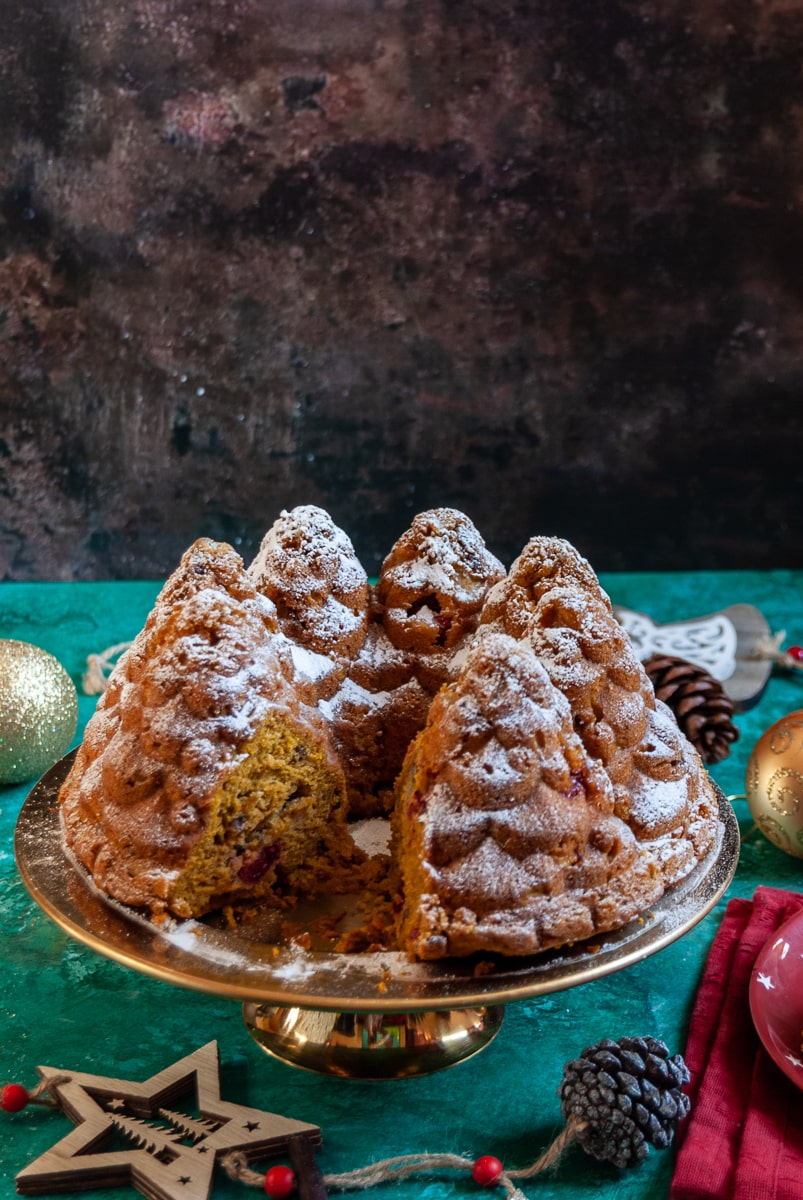 A pumpkin bundt cake in the shape of a fir trees on a gold cake stand, surrounded by christmas decorations on a brown and green backdrop