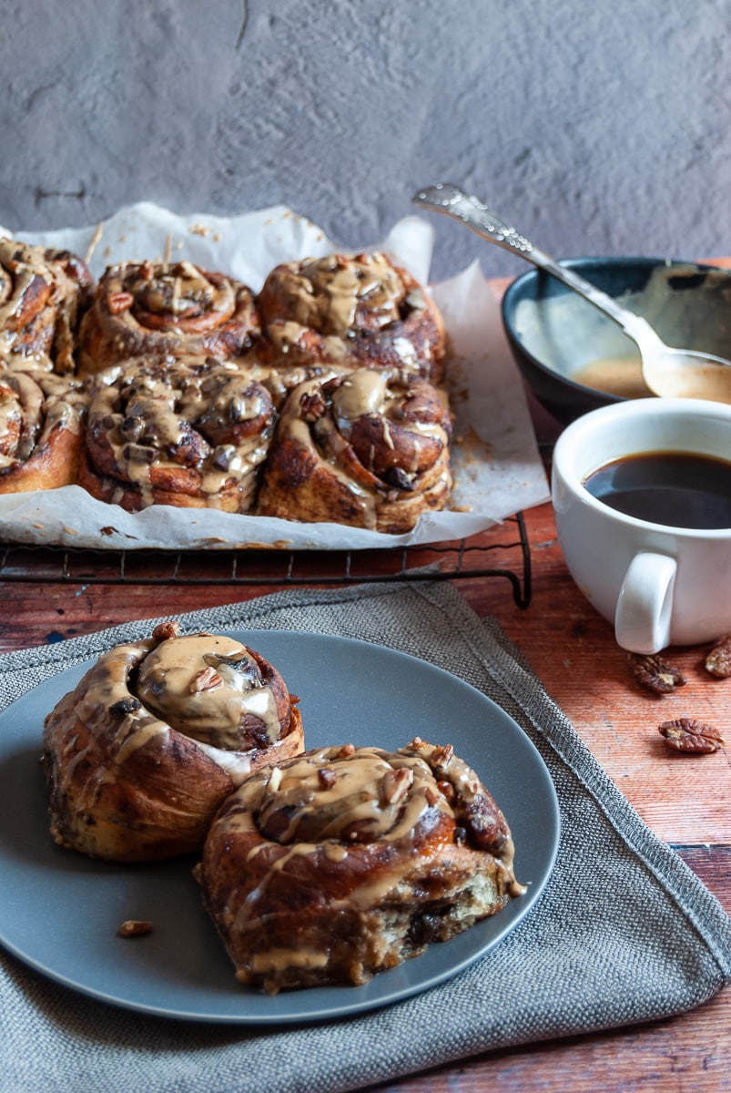 two sticky swirl buns covered in coffee icing and sprinkled with chopped pecans on a grey plate sitting on a grey linen napkin. More sticky buns on a wire rack in the background with a bowl of coffee icing with a silver spoon and a cup of coffee.