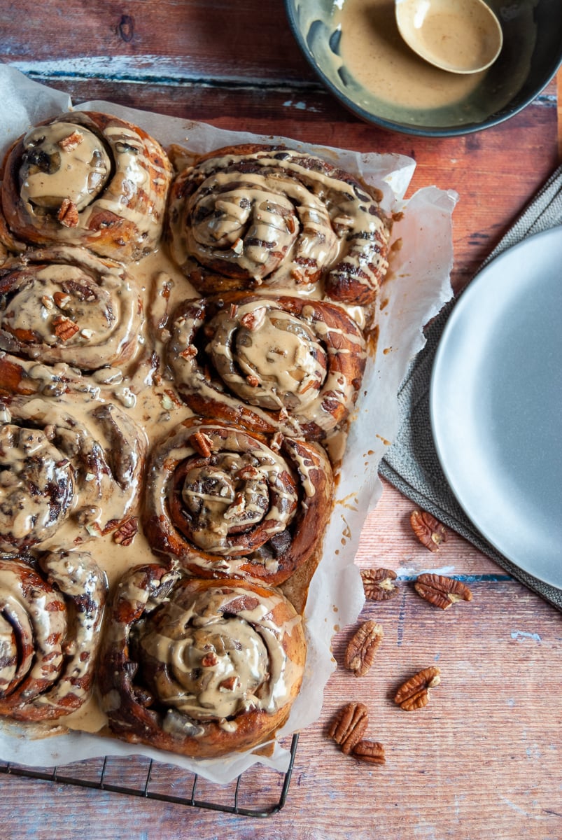 A batch of sticky buns covered in coffee icing and sprinkled with chopped pecans on a wooden background. A grey plate and napkin, a black bowl of coffee icing with a spoon can be partially seen.