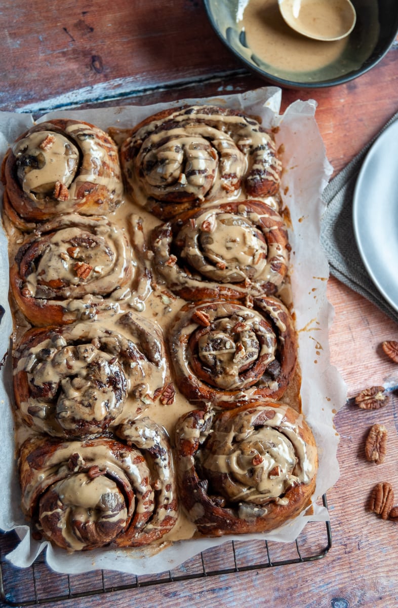 a batch of sticky buns covered in coffee icing and sprinkled with chopped pecans on a black wire rack and wooden table.