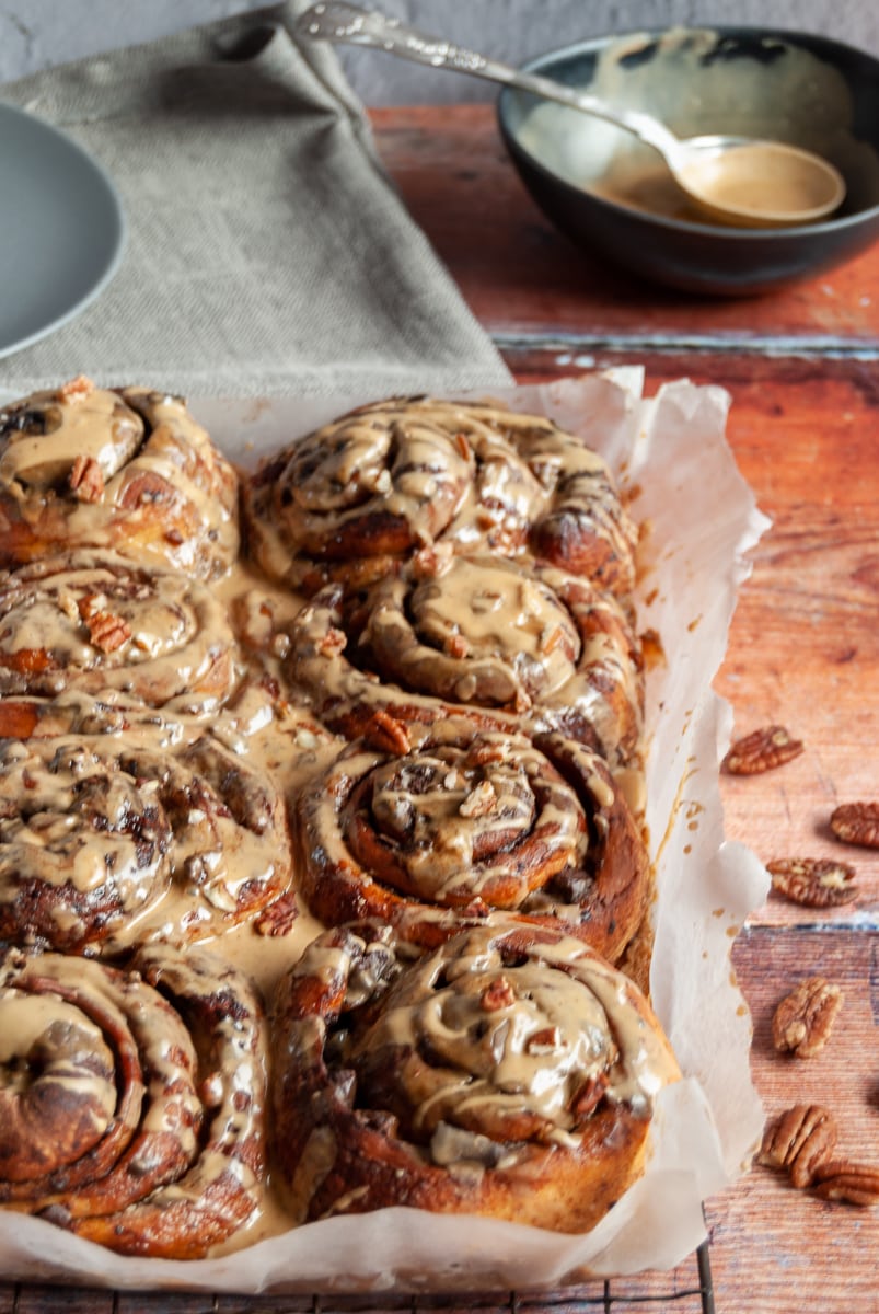 a batch of sticky buns covered in coffee icing and sprinkled with chopped pecans on a black wire rack, a black bowl of coffee icing and a grey napkin and plate are in the background