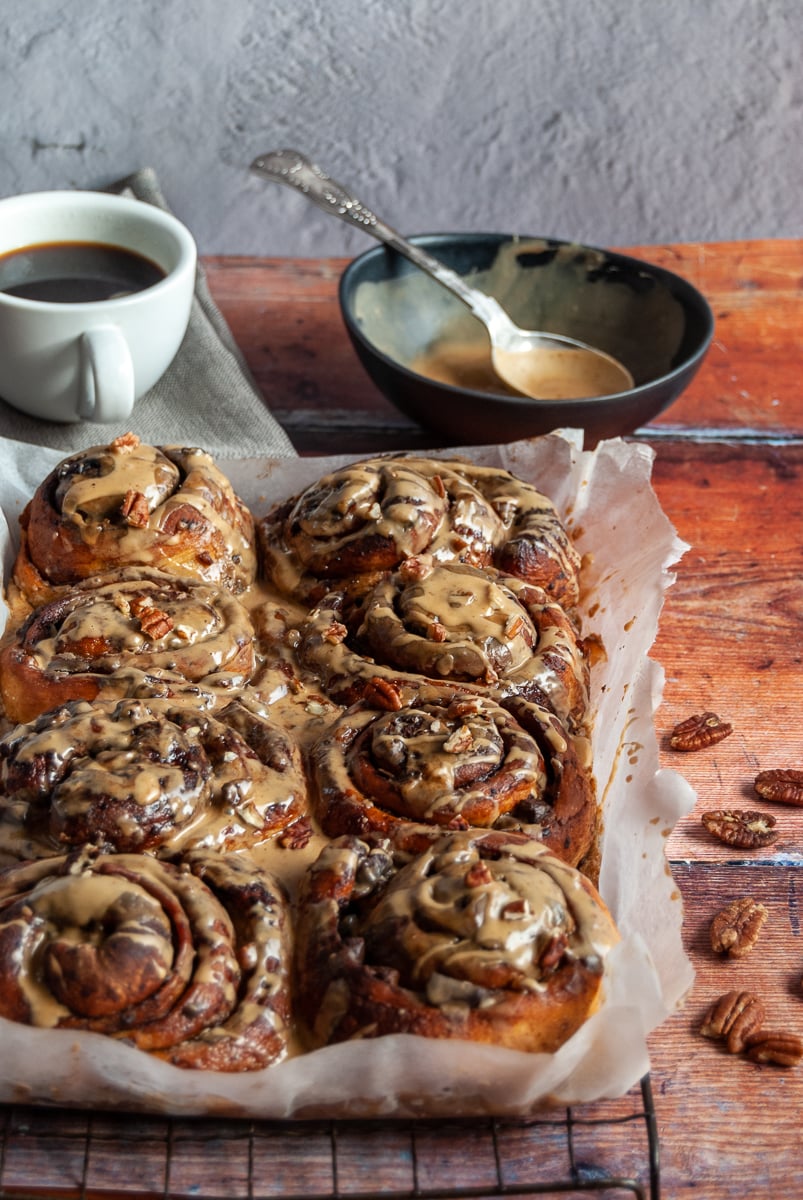 a batch of sticky buns covered in coffee icing and sprinkled with chopped pecans on a black wire rack, a small black bowl of coffee icing with a spoon and a white cup of coffee with a grey napkin.