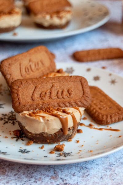 a mini cheesecake topped with melted Biscoff spread and a Lotus Biscuit on a white and silver star plate. More mini cheesecakeson a white plate can be seen in the background