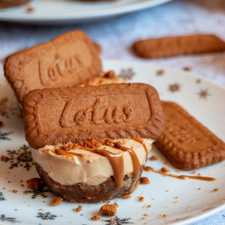 a mini cheesecake topped with melted Biscoff spread and a Lotus Biscuit on a white and silver star plate. More mini cheesecakeson a white plate can be seen in the background