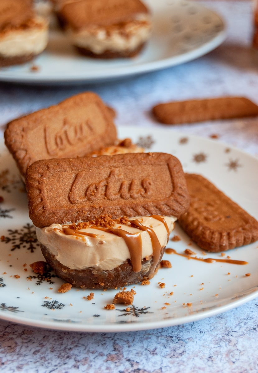 a mini cheesecake topped with melted Biscoff spread and a Lotus Biscuit on a white and silver star plate. More mini cheesecakeson a white plate can be seen in the background