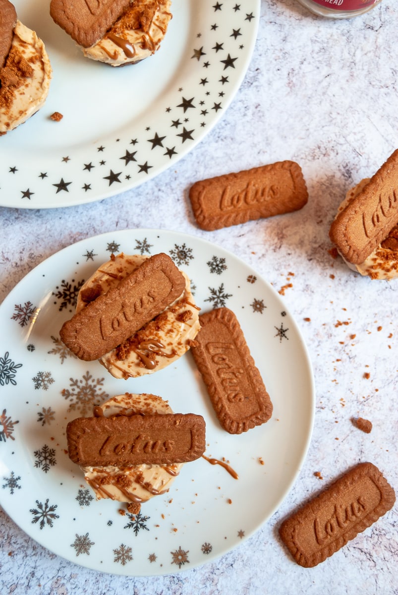 a flat lay photo of mini biscoff cheesecakes topped with a biscoff cookie on white and silver star plates. Crumbled Biscoff cookies are placed around the plates on a grey and white table top.