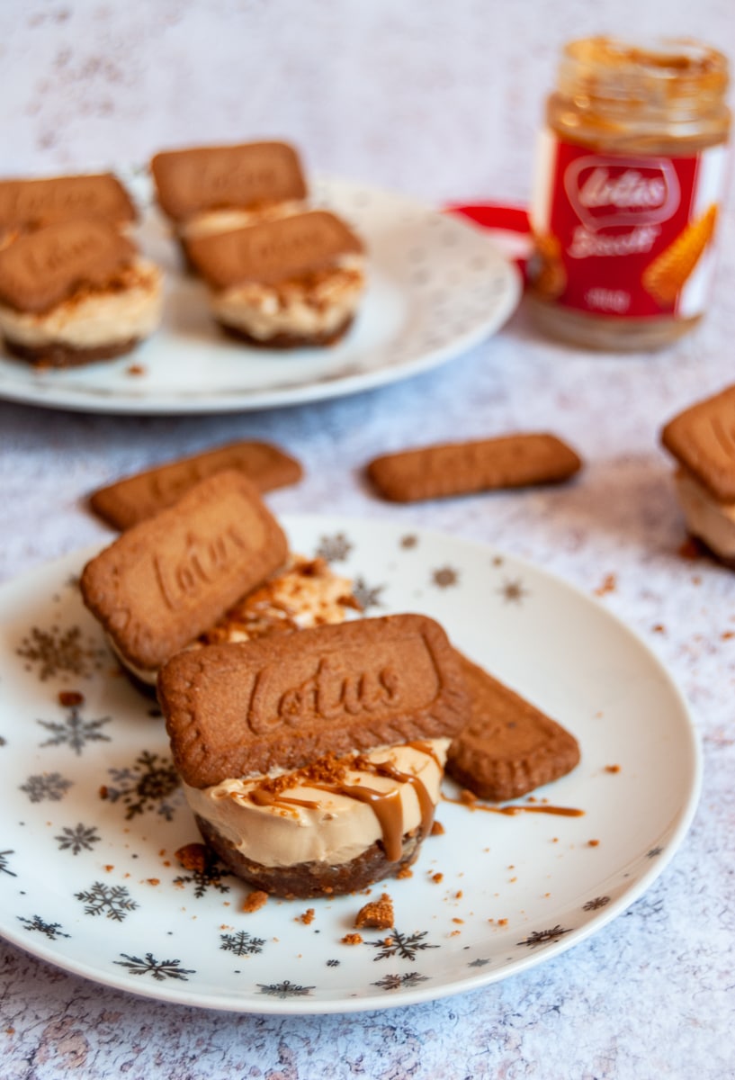 Mini cheesecake drizzled with melted Biscoff spread and topped with a Biscoff biscuit on a white and silver star plate. More Biscoff cheesecakes can be seen in the background along with a jar of Biscoff spread and more Biscoff biscuits.