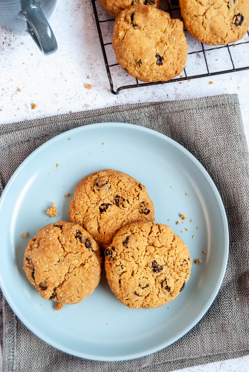 Three oatmeal raisin cookies on a light blue plate and grey napkin. More cookies on a wire rack can be partially seen.