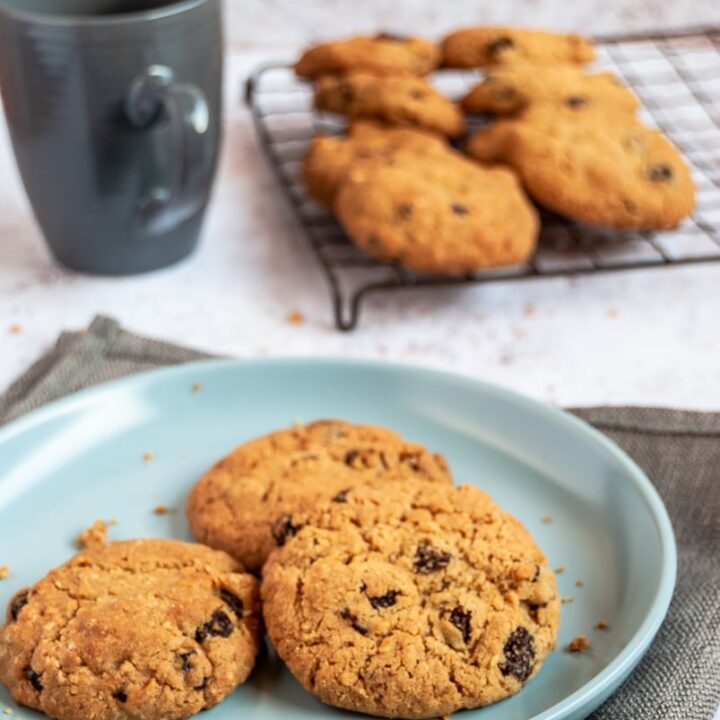 Three oat raisin cookies on a light blue plate, a navy coffee cup and a wire rack with more cookies.