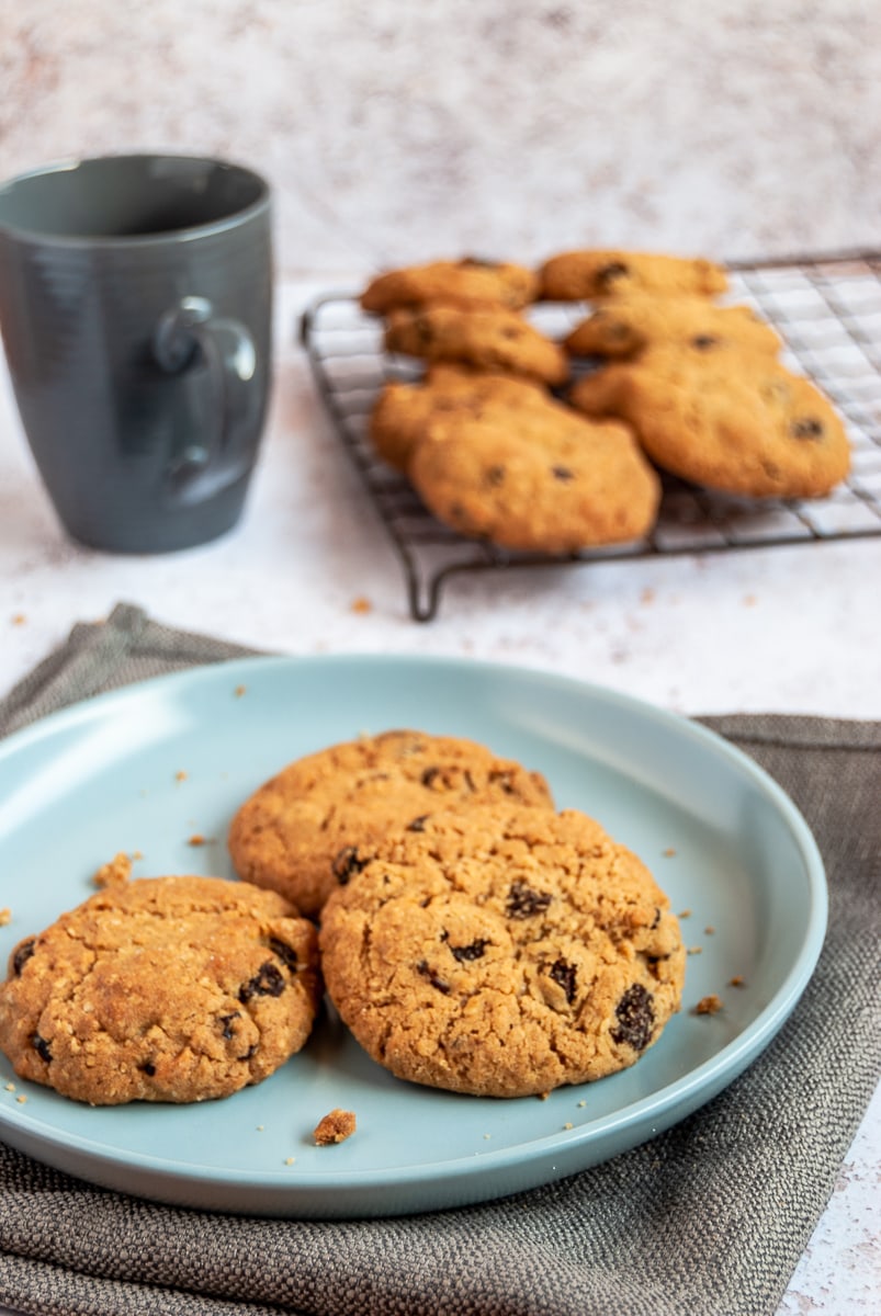 Three oat raisin cookies on a light blue plate, a navy coffee cup and a wire rack with more cookies.