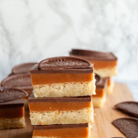 three square of millionaires shortbread stacked on top of each other, topped with a square of chocolate orange on a wooden board