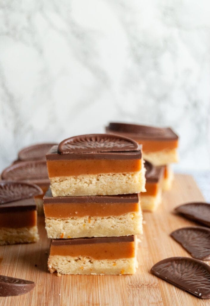 three squares of millionaires shortbread stacked on top of each other, topped with a square of chocolate orange on a wooden board