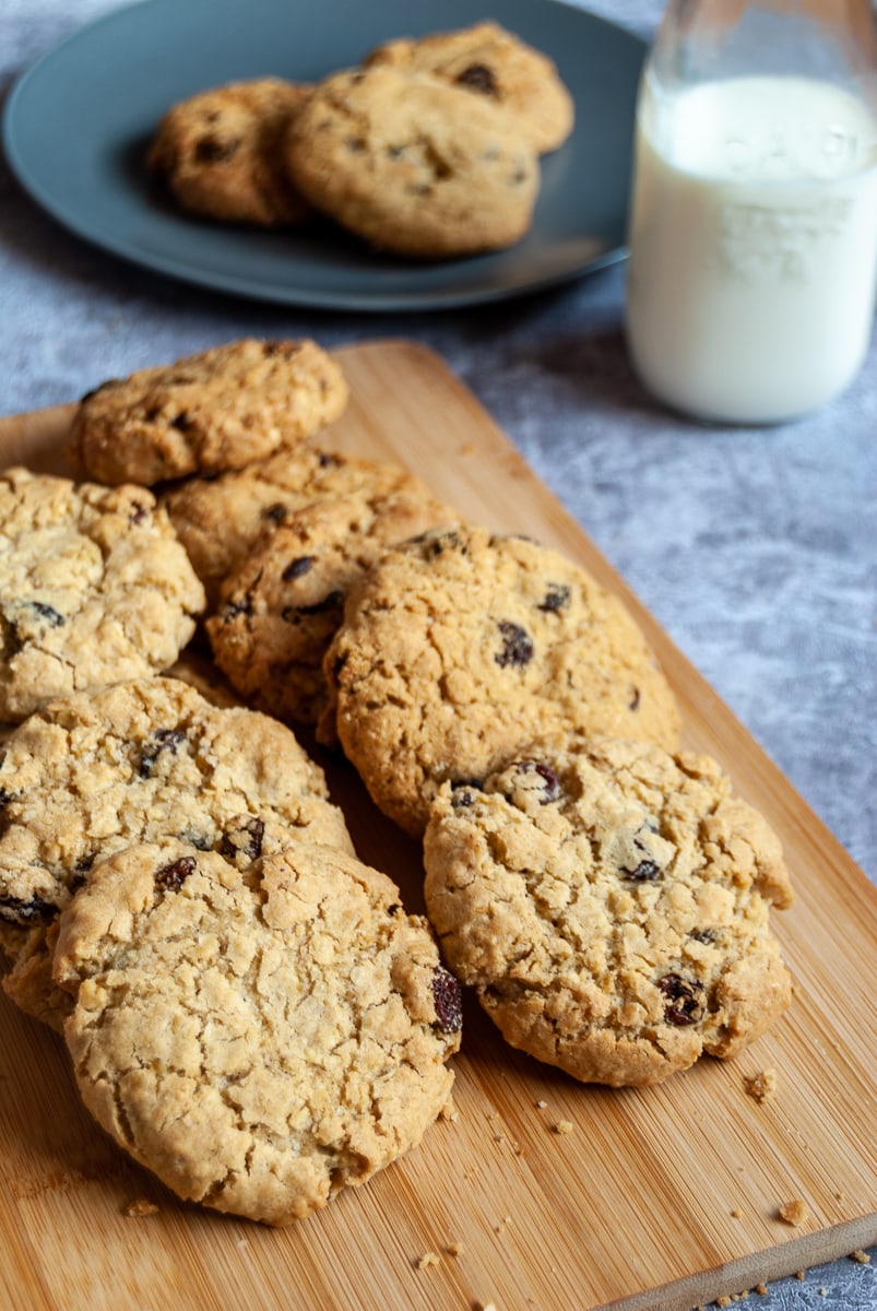 a batch of oatmeal raisin cookies on a wooden board, a blue plate with three cookies and a small glass bottle of milk