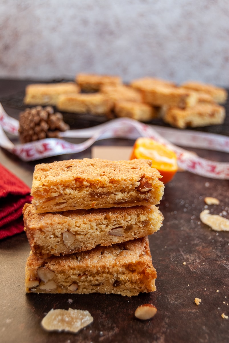 orange and almond cookie bars stacked on top of each other with a red napkin, red and white Christmas ribbon, a small orange and a pine cone. More cookie bars on a wire rack are in the background.