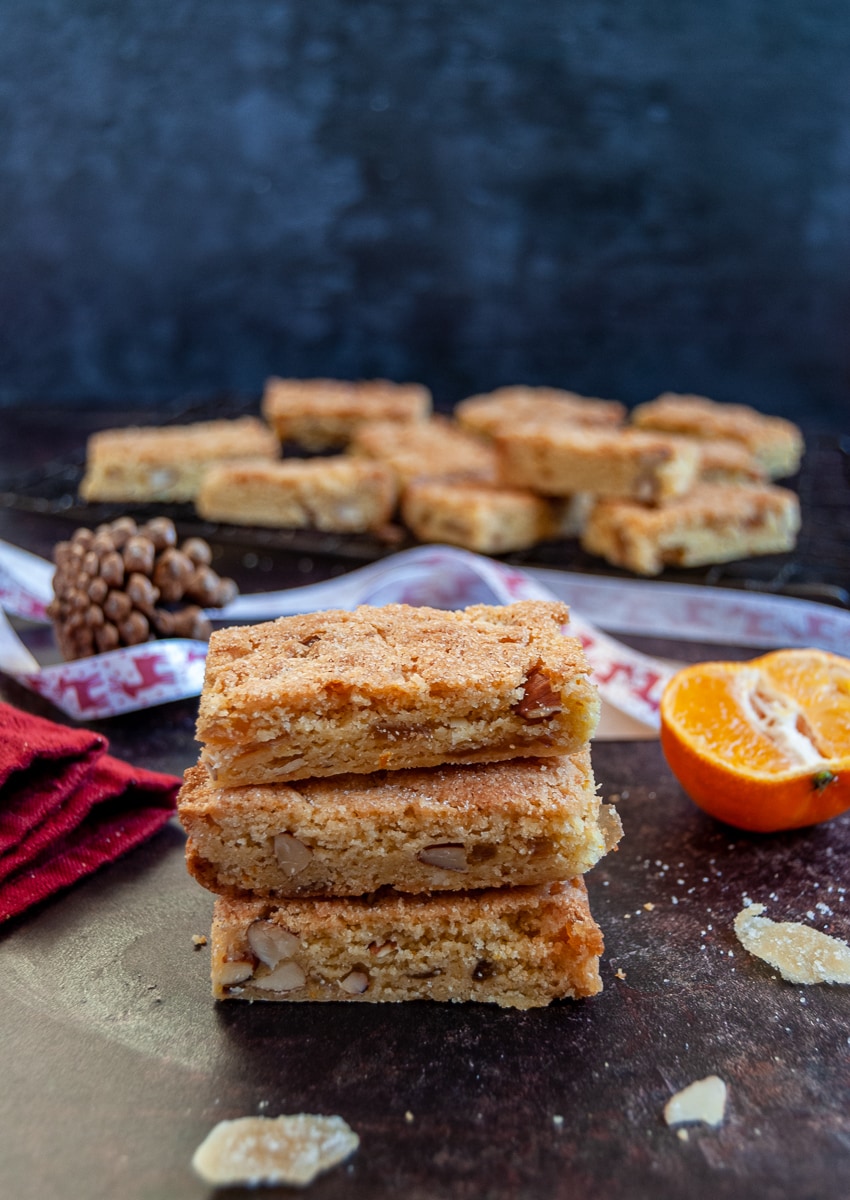 three orange and almond cookie bars stacked on top of each other, surrounded by red and white Christmas ribbon, a pine cone, a red napkin and a small orange. More of the bars can be seen in the background on a black wire rack