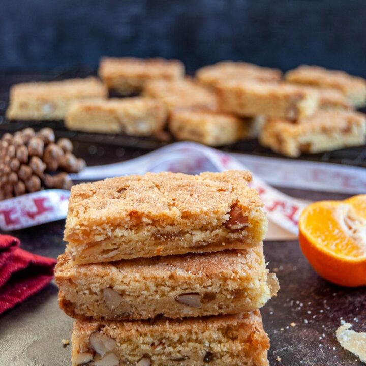 three orange almond bars stacked on top of each other, Christmas decorations and ribbons in the background and more of the orange bars on a black wire rack.