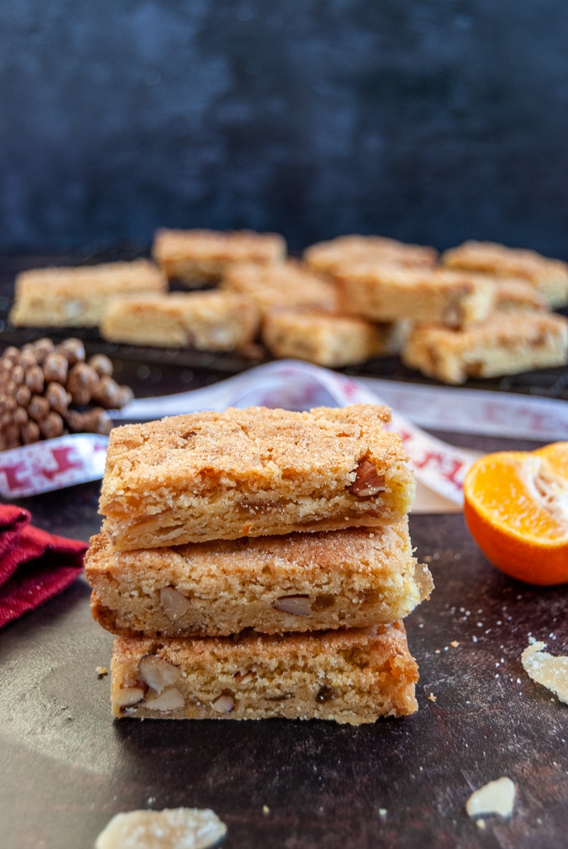 three orange almond bars stacked on top of each other, Christmas decorations and ribbons in the background and more of the orange bars on a black wire rack.