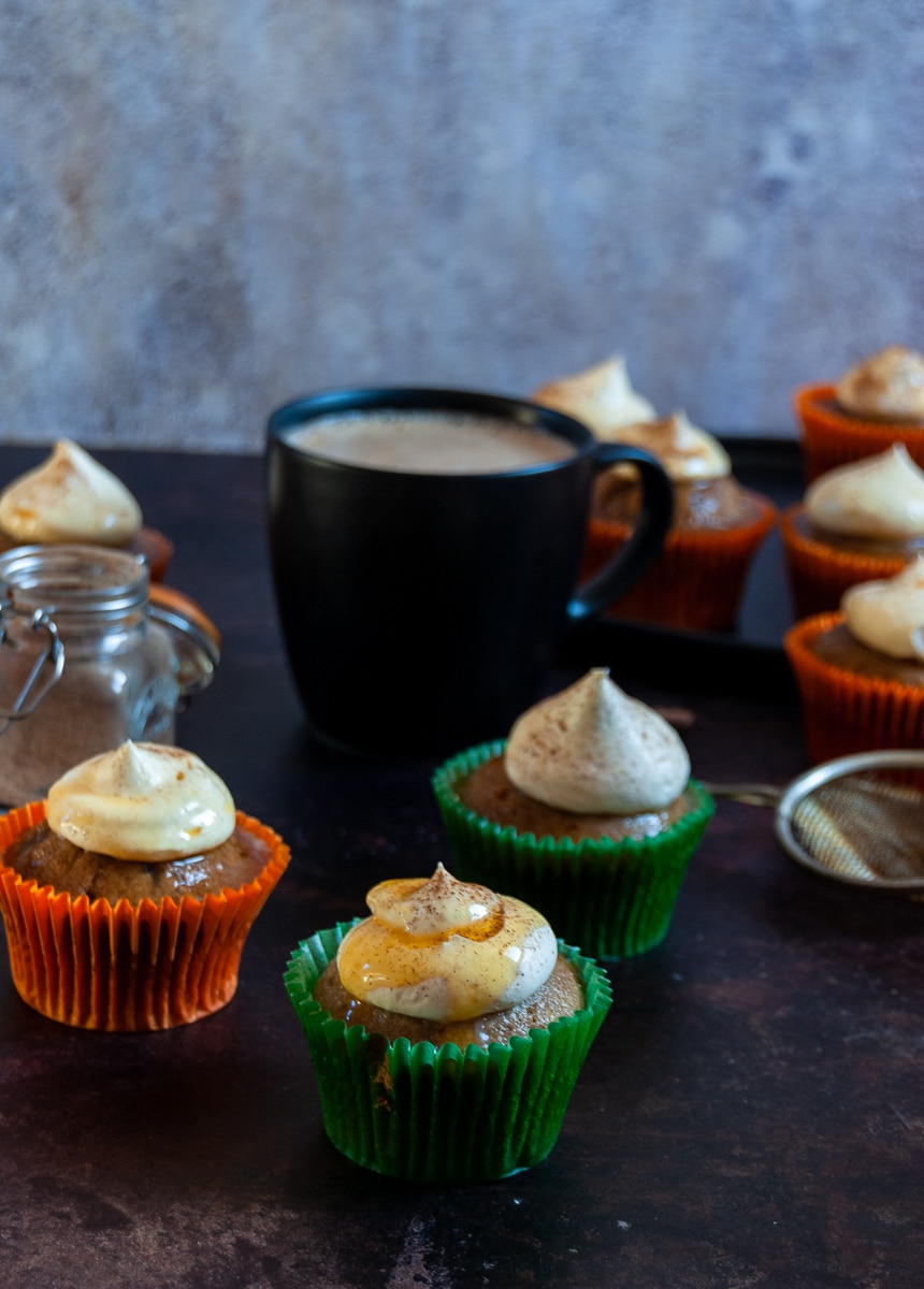 A pumpkin spice cupcake in a green cupcake liner topped with whipped cream and drizzled with caramel sauce. More cupcakes in the background with a black mug of coffee and a small jar of pumpkin spice 