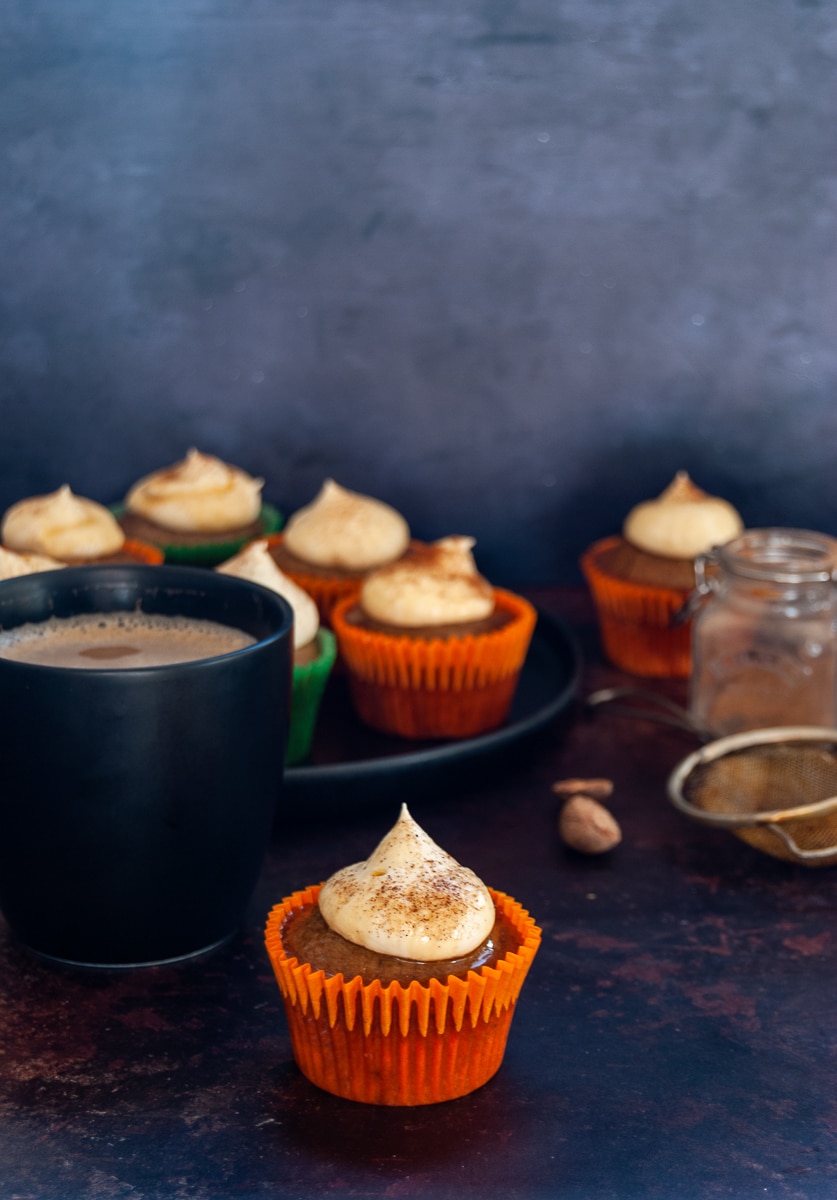 A pumpkin spice cupcake in an orange cupcake case topped with whipped cream and cinnamon, a black mug of coffee and more cupcakes in the background on a black plate and a small jar of pumpkin spice and a small rusted tea strainer