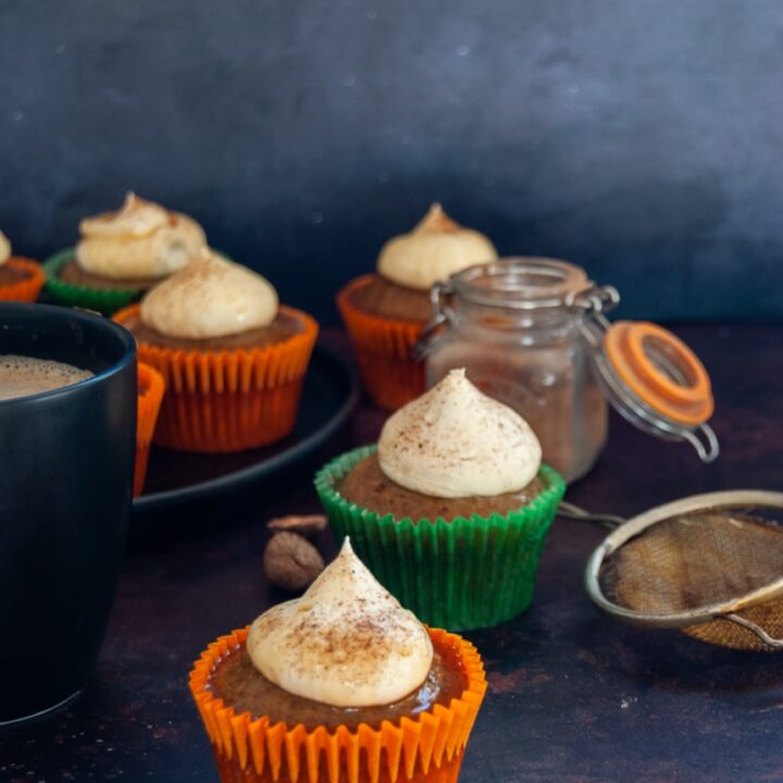 A pumpkin spice cupcake in an orange cupcake liner topped with whipped cream and dusted with cinnamon, more cupcakes on a black plate and a mug of coffee, a small pot of pumpkin spice and an icing sugar duster in the background.