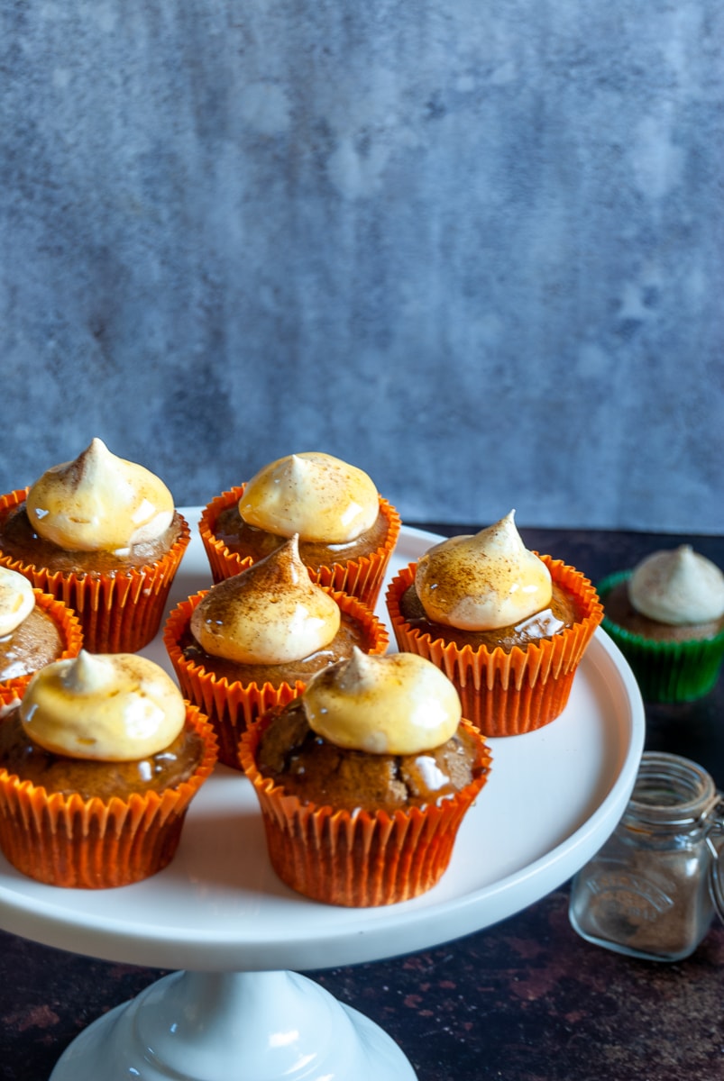 Pumpkin spice cupcakes in orange cupcake liners on a white cake stand, a small pot of pumpkin spice and more cupcakes in the background 