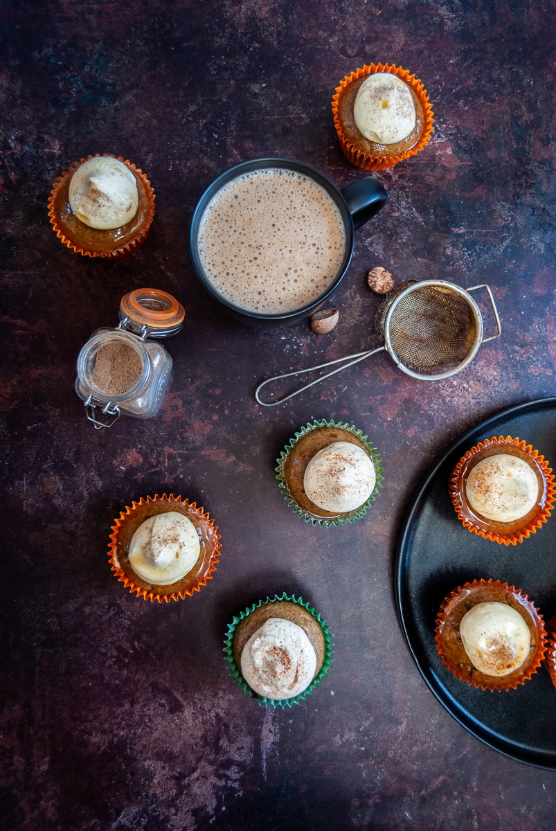 An overhead shot of pumpkin spice cupcakes topped with whipped cream and dusted with cinnamon, a little jar of pumpkin spice and a mug of coffee on an Autumn coloured backdrop 