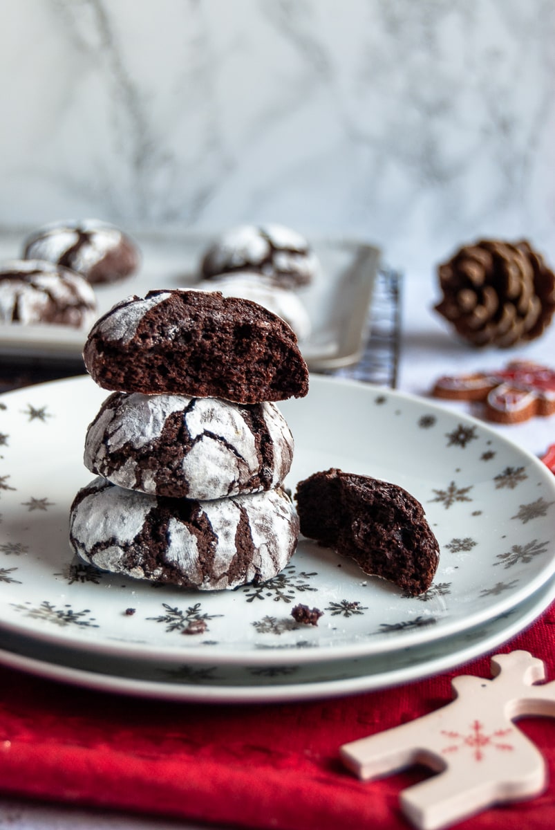 chocolate cookies dusted with confectioner's sugar on a white and silver star plate, a red napkin, a white reindeer decoration and more cookies on a baking sheet in the background.