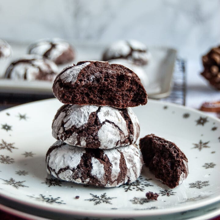 Chocolate cookies dusted with icing sugar on a white and silver star plate with a red napkin. Christmas decorations and more cookies on a baking sheet in the background