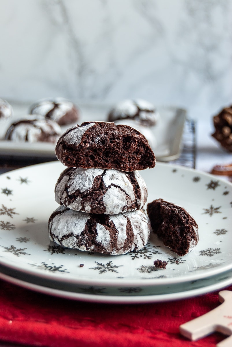 Chocolate cookies dusted with icing sugar on a white and silver star plate with a red napkin. Christmas decorations and more cookies on a baking sheet in the background